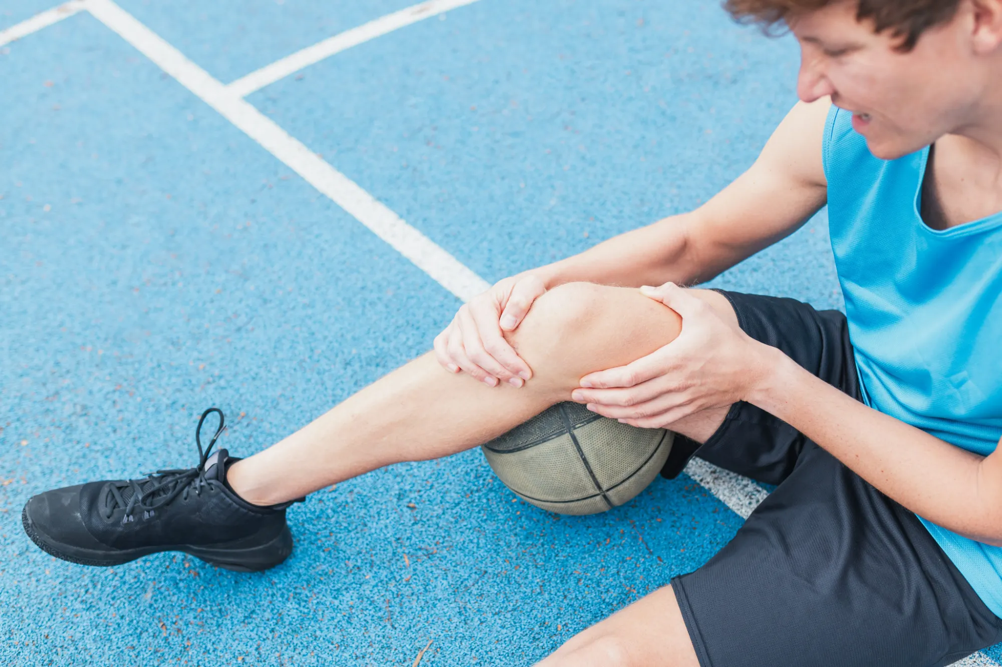 Young athlete sitting on a basketball court holding his knee after a possible ACL tear